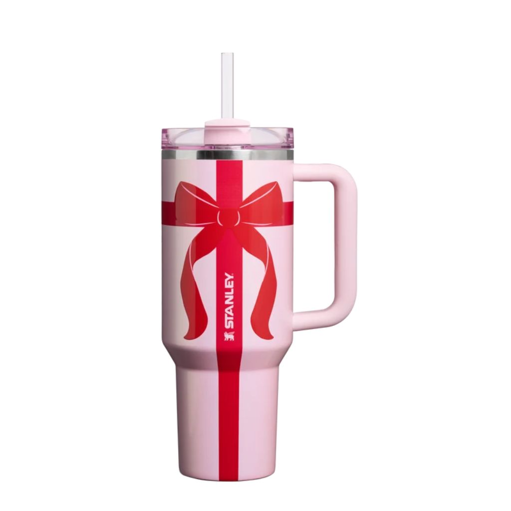 Stanley Soft Pink Ribbon tumbler featuring a pastel ballet-pink finish, oversized cherry-red gift-wrap bow with flowing ribbon tails, vertical red ribbon stripe, matching light pink handle, translucent pink lid, white straw, and white Stanley logo.